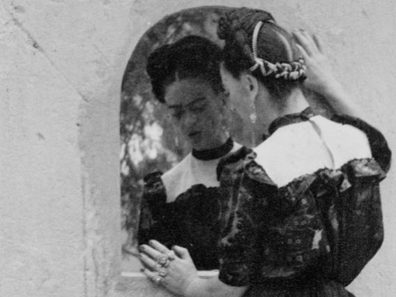 Image of Frida Kahlo standing and looking into a small mirror. Two hairless dogs stand at her feet.